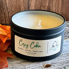 Load image into Gallery viewer, Candle in a tin labeled 'Cozy Cabin' on a wooden surface with fall leaves.